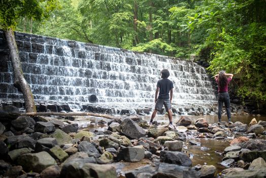 Water cascades over the tiered waterfall with lush woodland foliage behind. Two young men, African-American and Caucasion, stand on stones at base of falls.