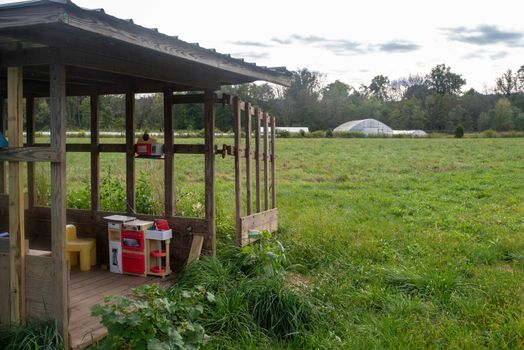 Rustic clubhouse with toys sits in green agricultural field on organic family farm with rural landscape and greenhouses in the background.