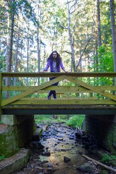 Caucasion man with long dark hair stands alone on a wooden bridge over a woodland creek reflecting the colorful surroundings.