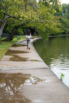One long haired young man walks away down a path by a pond. Sad puddles and a tree branch hangs overhead.