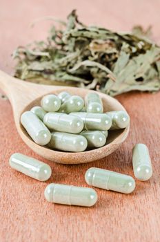 Herb medical capsules in wooden spoon with dry leaves herb on wooden background.