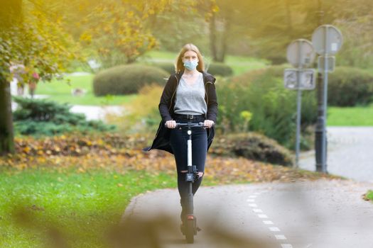 Casual caucasian teenager wearing protective face mask riding urban electric scooter in city park during covid pandemic. Urban mobility concept.