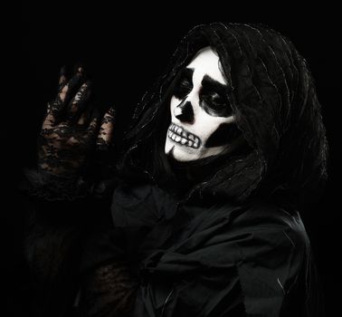 woman with a make-up skeleton stands in black clothes and a transparent hood, hands raised up, black background