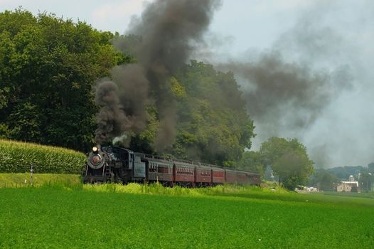 Antique Steam Passenger Train Puffing Lots of Black Smoke along Amish Countryside with Green Fields