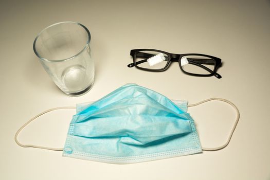 Surgical mask lying on the counter, glasses and an empty glass, top view