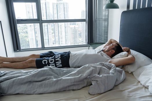 The guy is lying on the bed near the window with excellent breathtaking views of the city from a high floor.