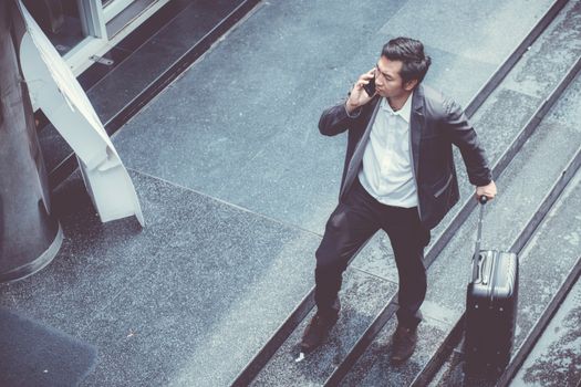 Asian elegant businessman wear suit talking mobile phone while walking holding suitcase outdoor on street, young man contact with customer appointment, communication business concept.