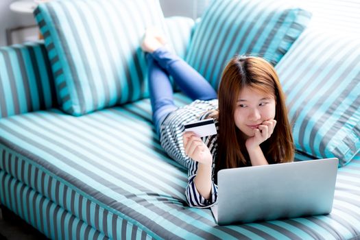 Beautiful of portrait young asian woman lying users credit card with laptop, Content girl shopping online and payment with notebook computer on sofa, lifestyle concept.
