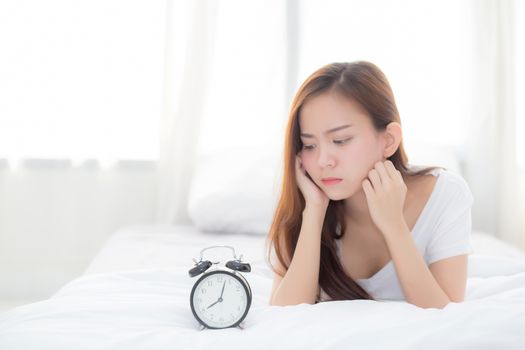 Beautiful of portrait young asian woman wake up in morning annoyed looking  alarm clock, girl lying hurry wake late with appointment with curtain background on bedroom, lifestyle concept.