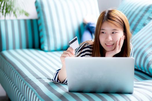 Beautiful of portrait young asian woman lying users credit card with laptop, Content girl shopping online and payment with notebook computer on sofa, lifestyle concept.