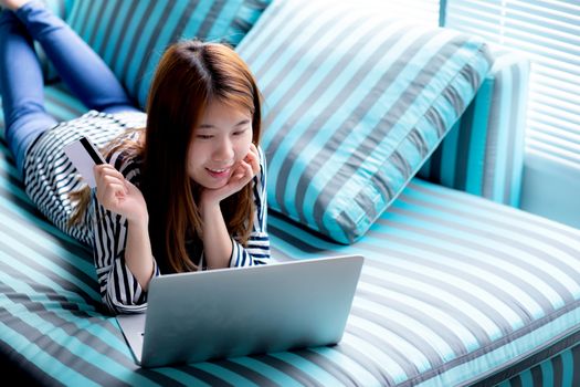 Beautiful of portrait young asian woman lying users credit card with laptop, Content girl shopping online and payment with notebook computer on sofa, lifestyle concept.