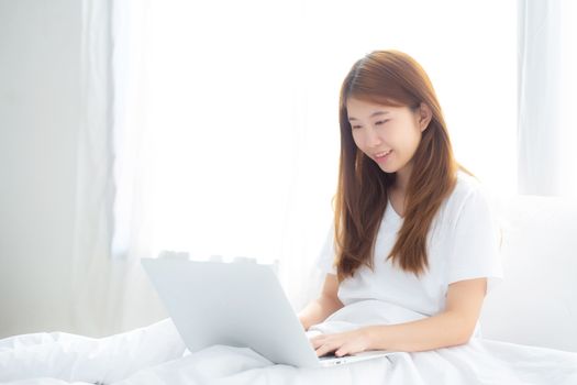 Portrait of beautiful asian young woman setting on bed using laptop computer at bedroom for leisure and relax, freelance with girl working notebook, communication and lifestyle concept.