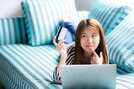 Beautiful of portrait young asian woman lying think use credit card with laptop, Content girl shopping online and payment with notebook computer on sofa, lifestyle concept.