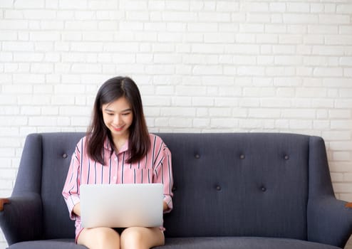 Beautiful of portrait asian young woman working online laptop sitting on sofa at living room, girl using notebook computer with connect to internet for distance job, business and success concept.