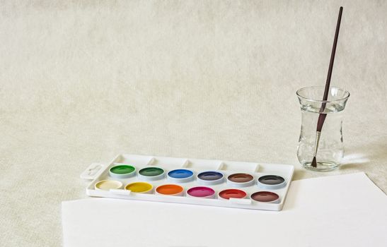 Watercolors, a glass of water with a brush and a sheet of white paper on a light background