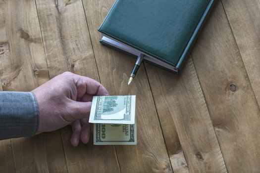 A $ 100 dollar bill is held by a man's hand near a diary with a fountain pen on a wooden surface.