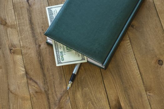 On the wooden surface is a diary with a pen and a part of the money bill is visible