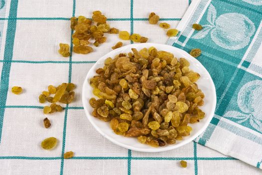 A white saucer with raisins stands on a cloth tablecloth and beside scattered raisins