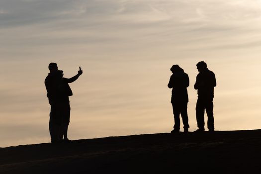 Silhouettes of tourists taking pictures at sunset in the Sahara desert Morocco. Set against a pale sky of blue and gold tourists on top of a sand dune. High quality photo