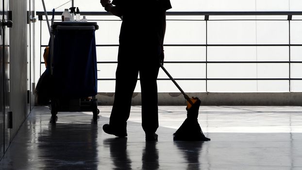 Silhouette image of cleaning service people sweeping floor with mop and other equipment on trolley.