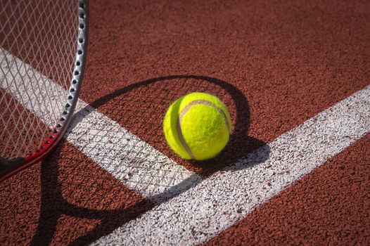 Tennis ball, line and racket on an outdoor court with the racket standing on end casting a shadow across the all weather surface