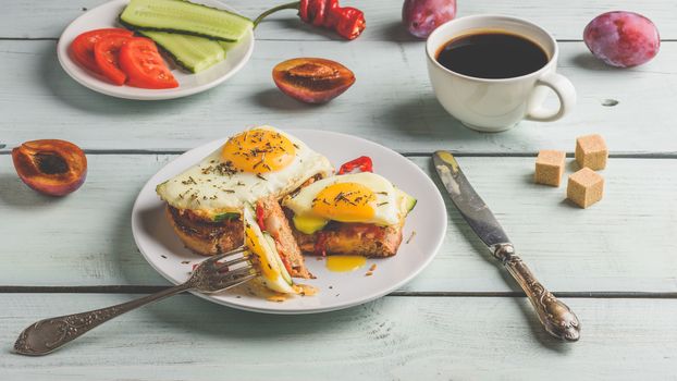 Sandwiches with vegetables and fried egg on white plate, cup of coffee and some fruits over wooden background.