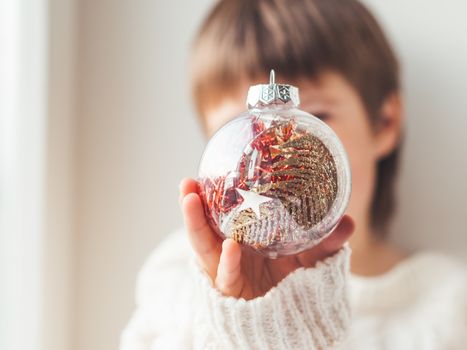 Kid with decorative ball for Christmas tree.Boy in cable-knit oversized sweater.Cozy outfit for snuggle weather.Transparent ball with red, golden spangles inside.Winter holiday spirit.New year.