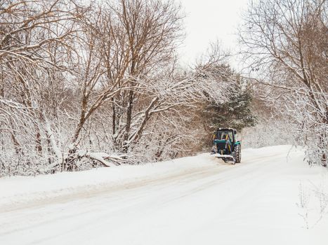 Snowplow clears rural road of snow debris after heavy snowstorm. Winter natural background with trees and country road under snow. Countryside landscape.