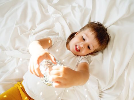 Little boy plays with sparkling mirror disco ball in bed and laughing happily. Joyful toddler. Playful child and sunny morning in cozy home.