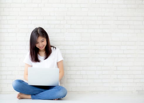 Beautiful of portrait asian young woman working online laptop sitting on floor brick cement background, freelance girl using notebook computer with connect to internet for distance job, business and lifestyle concept.