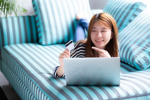 Beautiful of portrait young asian woman lying users credit card with laptop, Content girl shopping online and payment with notebook computer on sofa, lifestyle concept.