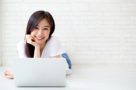 Beautiful of portrait asian young woman working online laptop lying on floor brick cement background, freelance girl using notebook computer with connect to internet for distance job, business and lifestyle concept.