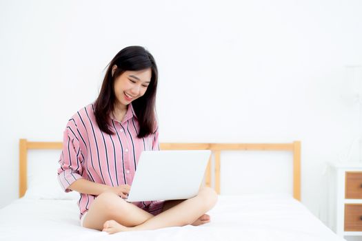 Portrait of beautiful asian young woman setting on bed using laptop computer at bedroom for leisure and relax, freelance with girl working notebook, communication and lifestyle concept.