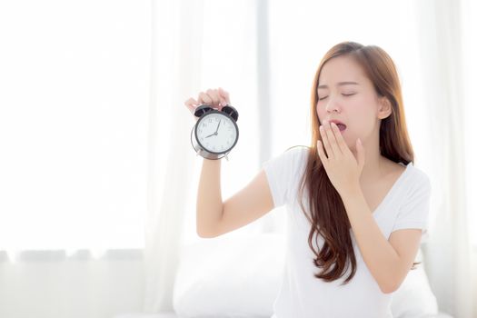 Beautiful young asian woman wake up in morning yawn and annoyed alarm clock holding hand, girl standing hurry wake late with appointment with curtain background on bedroom, lifestyle concept.
