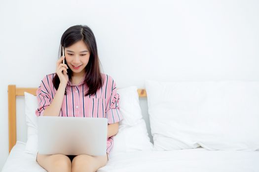 Portrait of beautiful asian young woman setting on bed using laptop computer and talk smart phone at bedroom for leisure and relax, freelance with girl working notebook and calling telephone, communication and lifestyle concept.