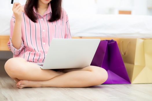 Closeup asian woman shopping online with laptop computer sitting on floor on room, girl holding credit card purchase and shopping bags, lifestyle concept.