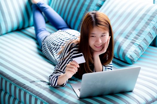 Beautiful of portrait young asian woman lying users credit card with laptop, Content girl shopping online and payment with notebook computer on sofa, lifestyle concept.