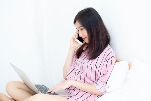 Portrait of beautiful asian young woman setting on bed using laptop computer and talk smart phone at bedroom for leisure and relax, freelance with girl working notebook and calling telephone, communication and lifestyle concept.