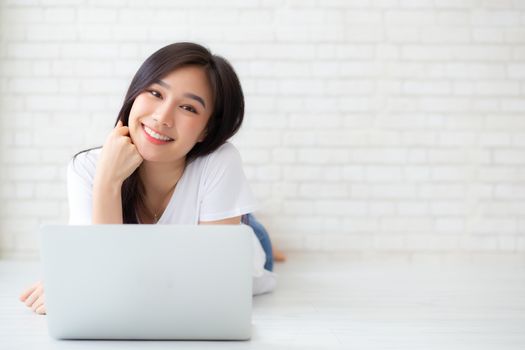 Beautiful of portrait asian young woman working online laptop lying on floor brick cement background, freelance girl using notebook computer with connect to internet for distance job, business and lifestyle concept.