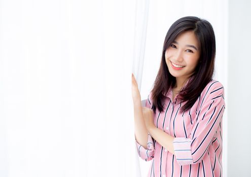 Portrait of beautiful young asian woman standing the window and smile while wake up with sunrise at morning, lifestyle and relax concept.