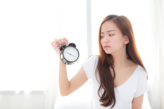 Beautiful young asian woman wake up in morning annoyed alarm clock holding hand, girl standing hurry wake late with appointment with curtain background on bedroom, lifestyle concept.