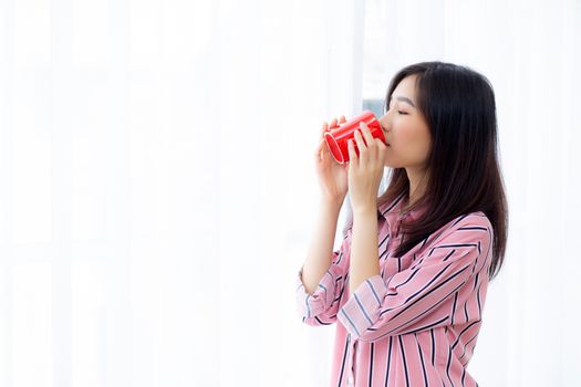 Beautiful of portrait young asian woman with drink a cup of coffee standing curtain window background in bedroom, girl relax in morning at home, lifestyle concept.