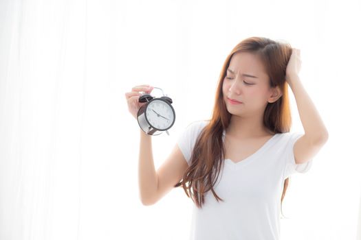 Beautiful young asian woman wake up in morning annoyed alarm clock holding hand, girl standing hurry wake late with appointment with curtain background on bedroom, lifestyle concept.