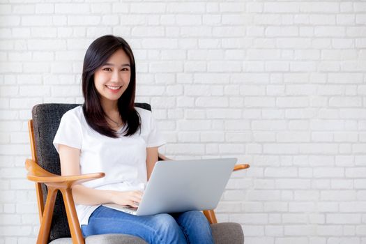 Beautiful of portrait asian young woman working online laptop sitting on chair on cement brick white background, girl using notebook computer, business concept.