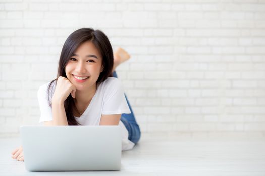 Beautiful of portrait asian young woman working online laptop lying on floor brick cement background, freelance girl using notebook computer with connect to internet for distance job, business and lifestyle concept.
