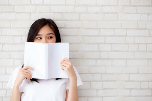 beautiful portrait young asian woman happy hiding behind open the book with cement or concrete background, girl standing reading for learning, education and knowledge concept.
