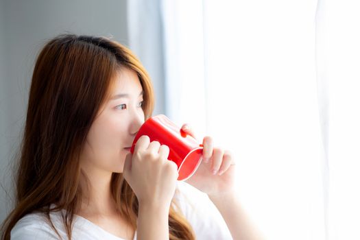 Beautiful of portrait young asian woman with drink acup of coffee standing curtain window background in bedroom, girl relax in morning at home, lifestyle concept.