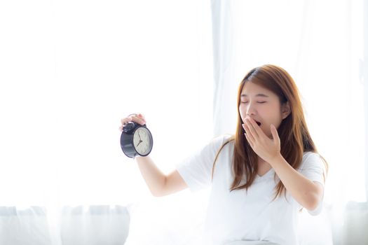 Beautiful young asian woman yawn wake up in morning annoyed alarm clock holding hand, standing hurry wake late with appointment with curtain background on bedroom, lifestyle concept.