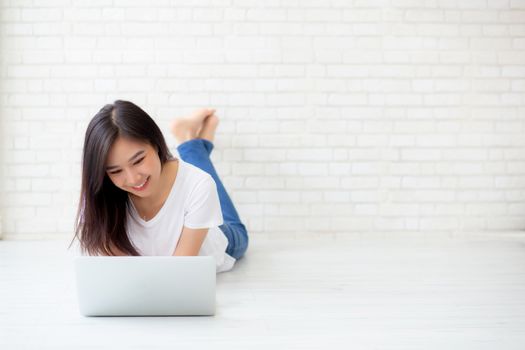 Beautiful of portrait asian young woman working online laptop lying on floor brick cement background, freelance girl using notebook computer with connect to internet for distance job, business and lifestyle concept.