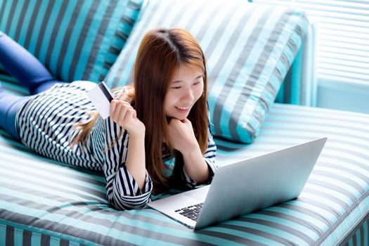 Beautiful of portrait young asian woman lying users credit card with laptop, Content girl shopping online and payment with notebook computer on sofa, lifestyle concept.